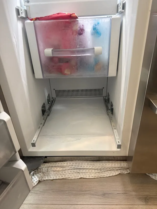 Freezer compartment with its lower basket removed, a layer of ice across its floor and towels on the floor below