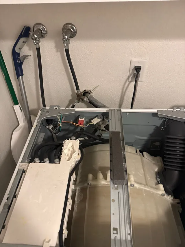 Front-load washer seen from above with its top panel removed, showing the tub, water valves and hoses