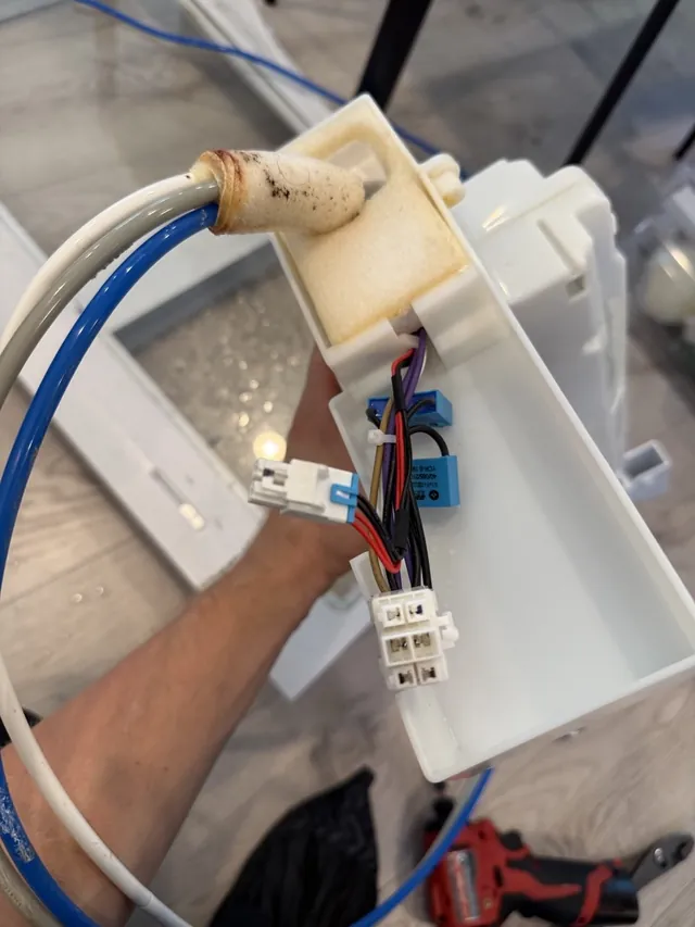 Hand holding a refrigerator ice and water assembly with its supply tubes and wiring connectors